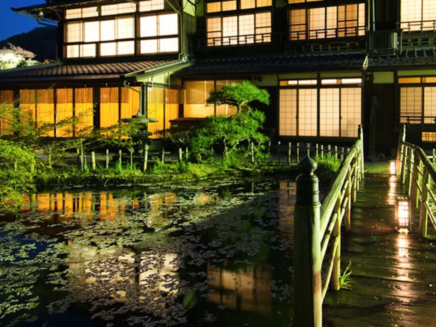 Ryokan traditionnel illuminé la nuit, son étang couvert de nénuphars et sa passerelle de bambou