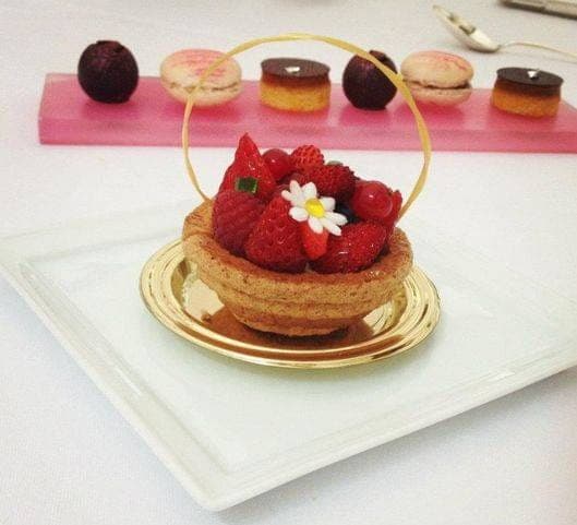 Tartelette aux fraises dressée sur un plat blanc, macarons et petits fours en arrière-plan
