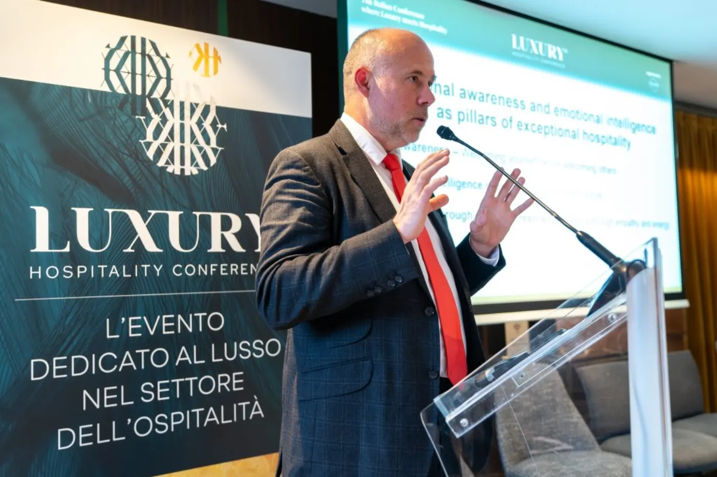 Orateur en costume gris s'exprimant au pupitre devant le kakémono italien de la Luxury Hospitality Conference
