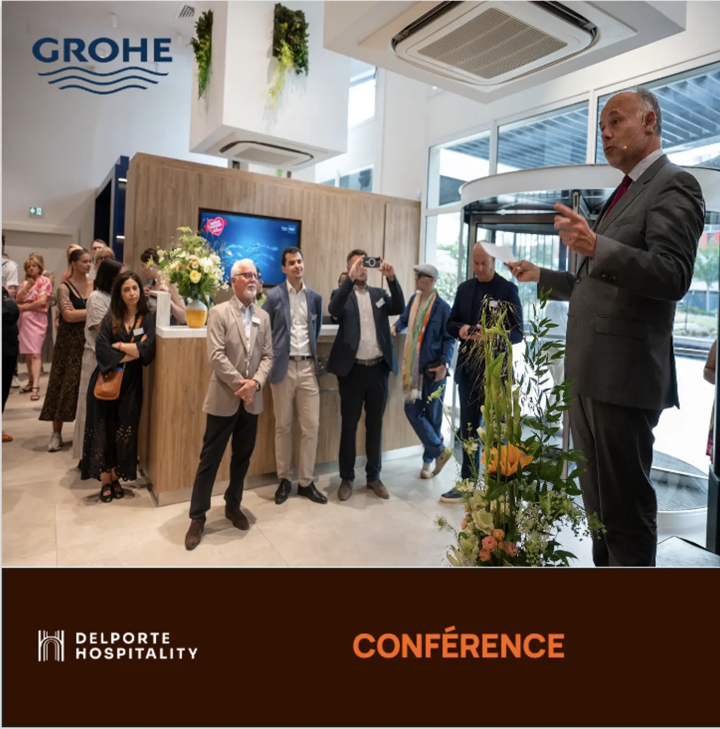 Conférencier en micro-casque s'adressant à un public debout dans un showroom GROHE, bandeau Delporte Hospitality