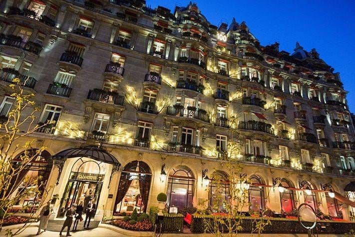 Façade du Plaza Athénée illuminée à la nuit tombée, ses balcons fleuris et son auvent d'entrée