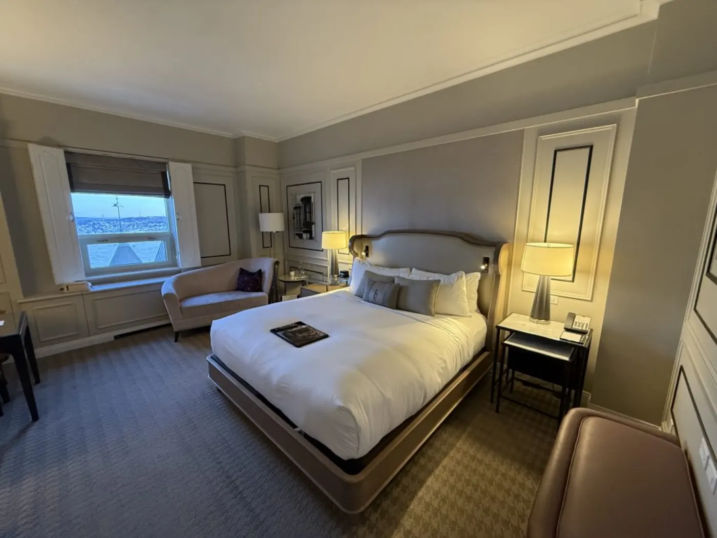 Chambre d'hôtel aux boiseries beiges, grand lit blanc, fauteuil près de la fenêtre et vue sur le fleuve