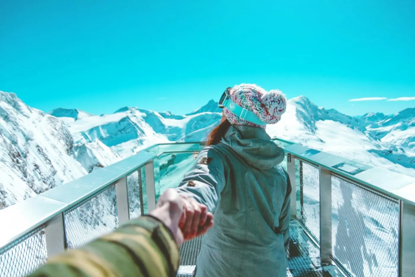 A woman in winter clothing holds a hand and walks on a scenic snow-covered mountain deck in Zell am See.