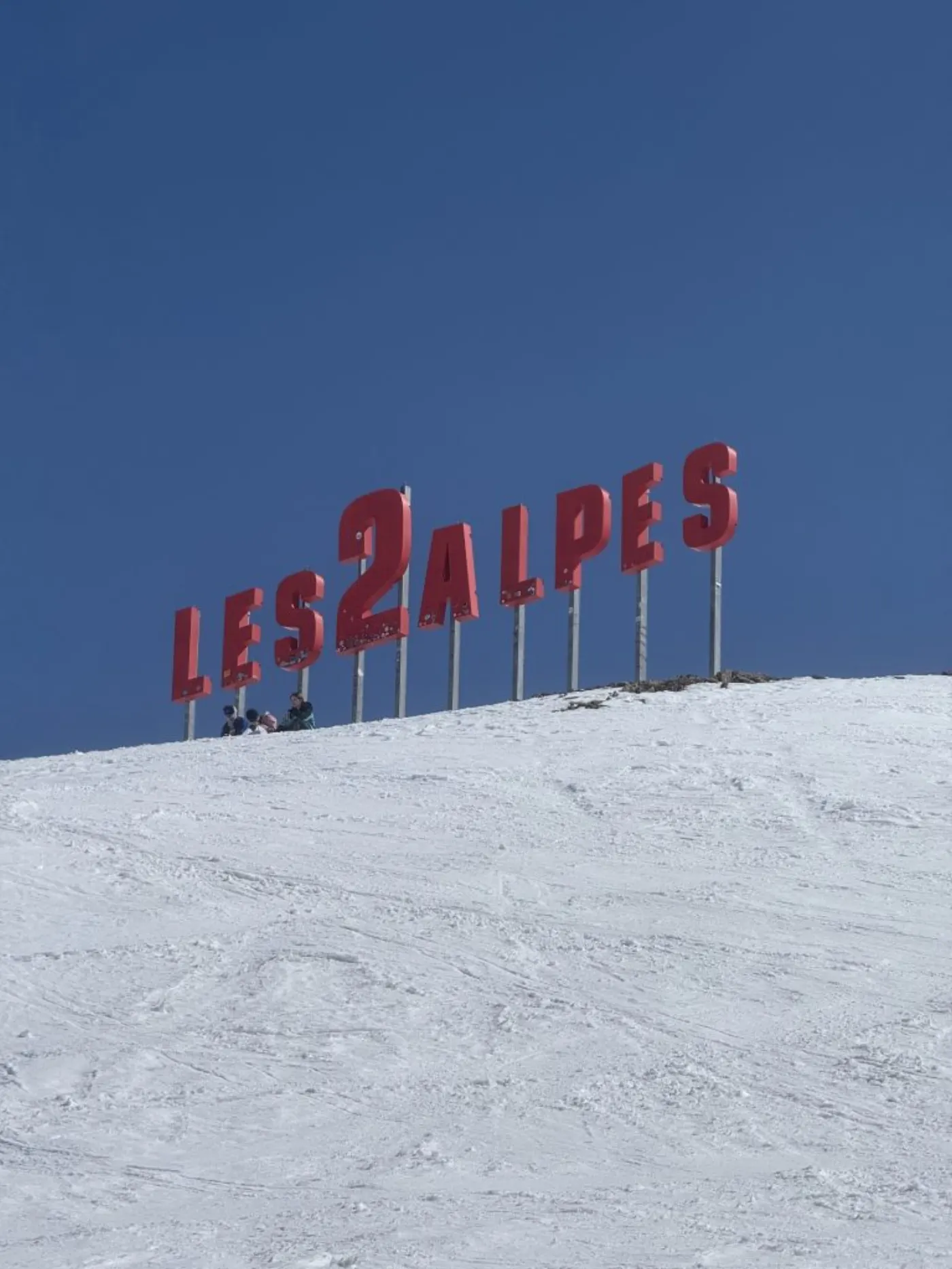 Lettres rouges Les 2 Alpes plantées en haut d'une piste enneigée sous un ciel bleu, quelques skieurs assis au pied
