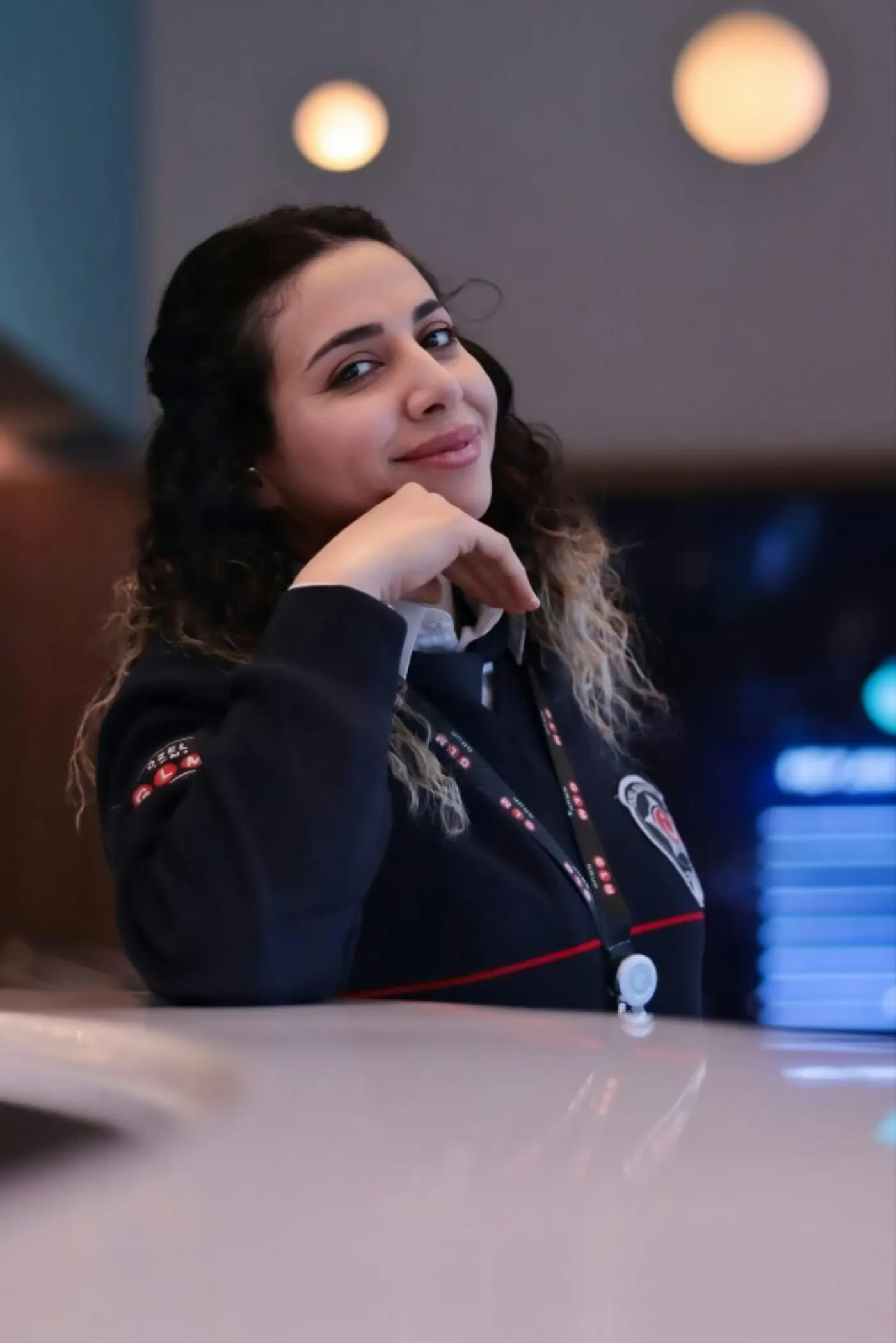 A confident woman in professional attire smiles warmly while leaning on a desk indoors.