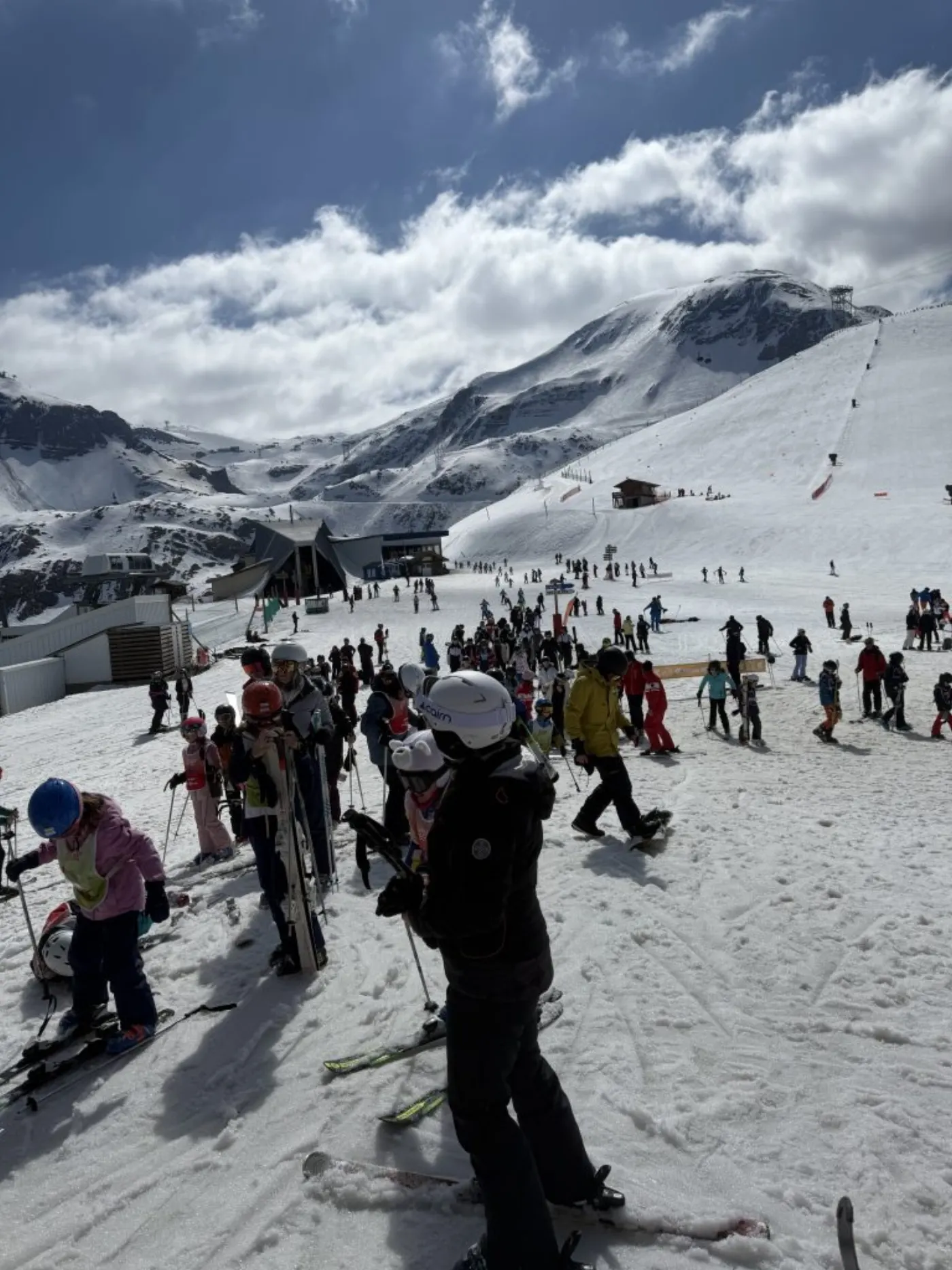Foule de skieurs et d'enfants en cours au pied des pistes, moniteurs en combinaison rouge et gare de remontée mécanique