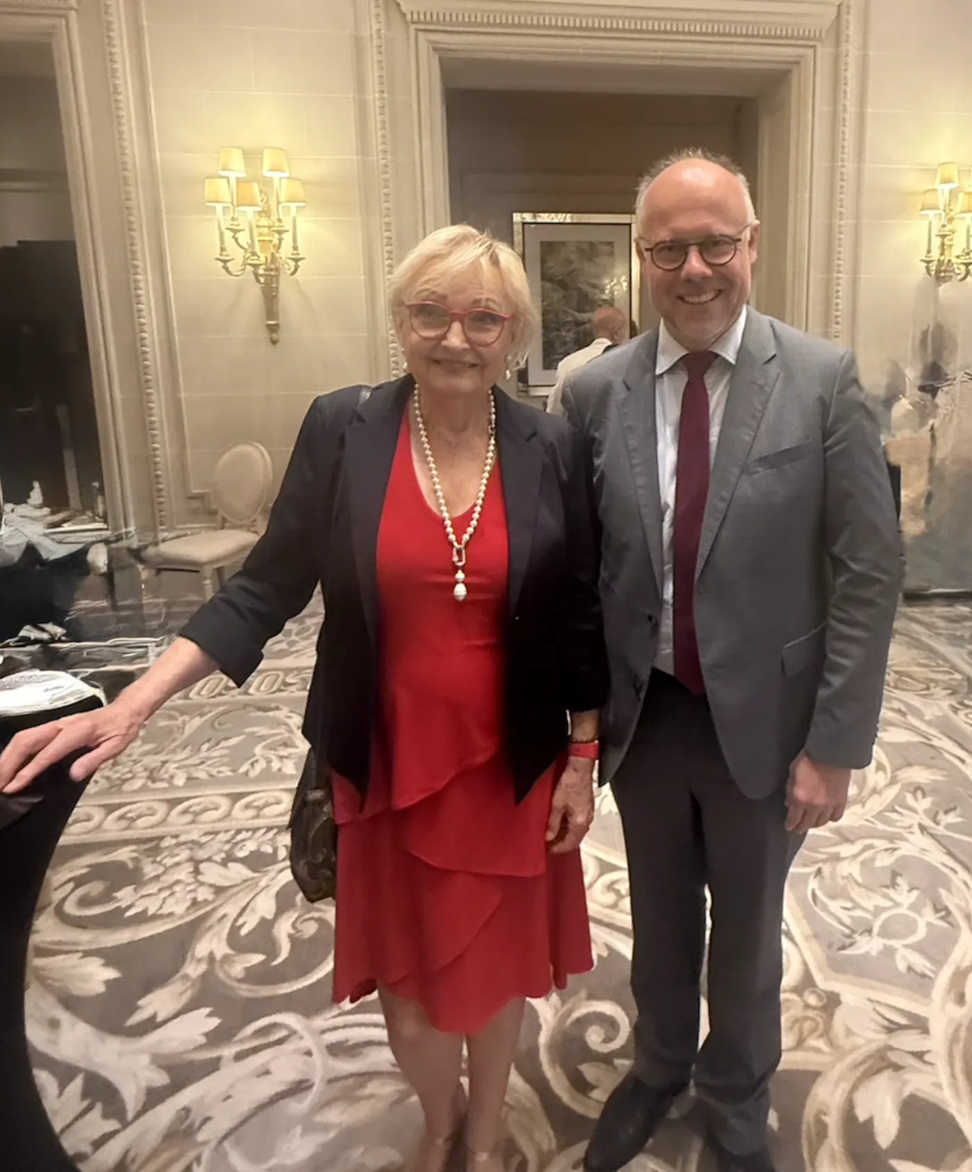 Deux invités posant côte à côte dans le salon d'un hôtel, femme en robe rouge et collier de perles, homme en costume gris