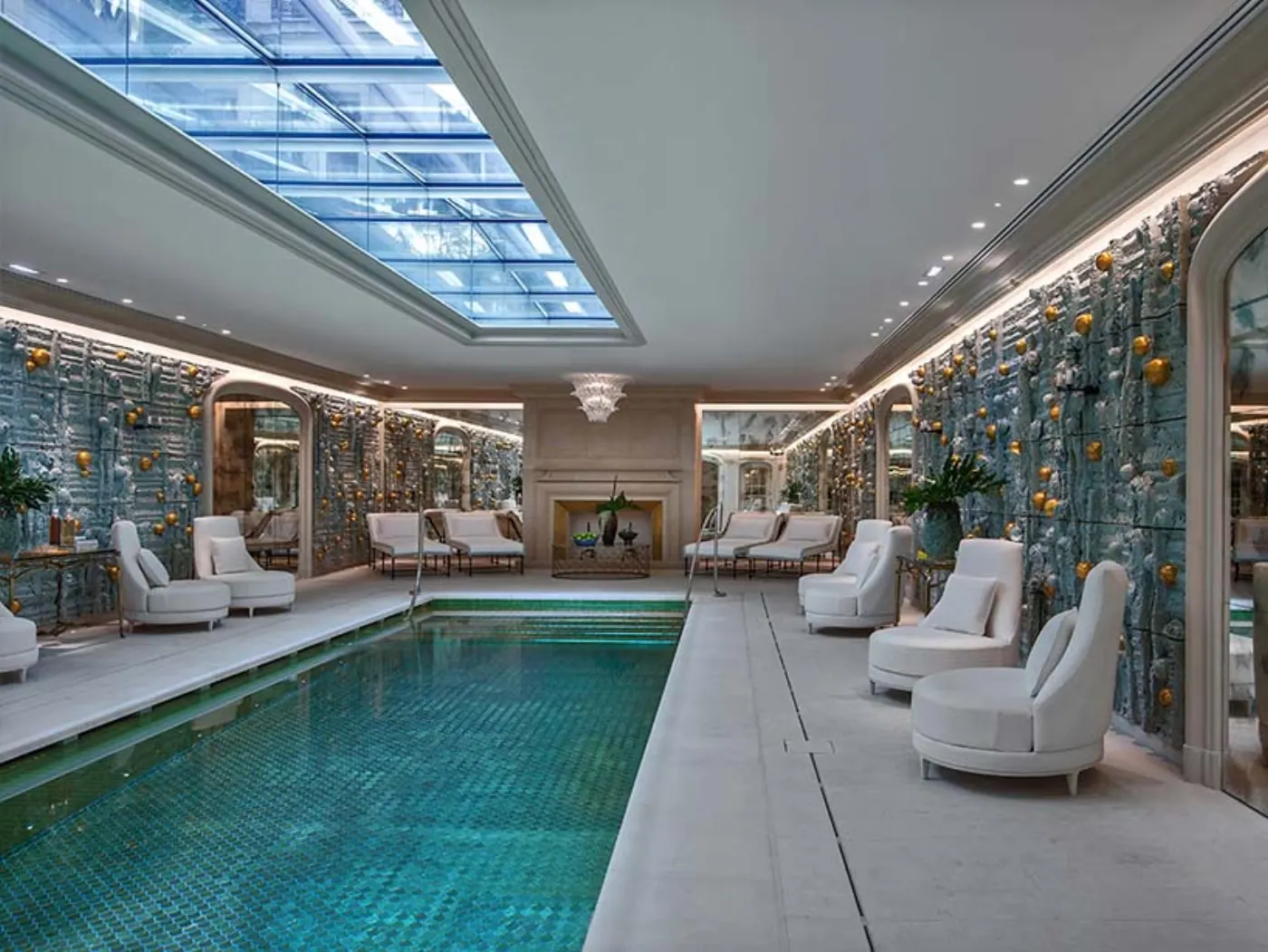 Piscine intérieure en mosaïque turquoise sous une verrière, fauteuils blancs et murs sculptés ponctués de sphères dorées