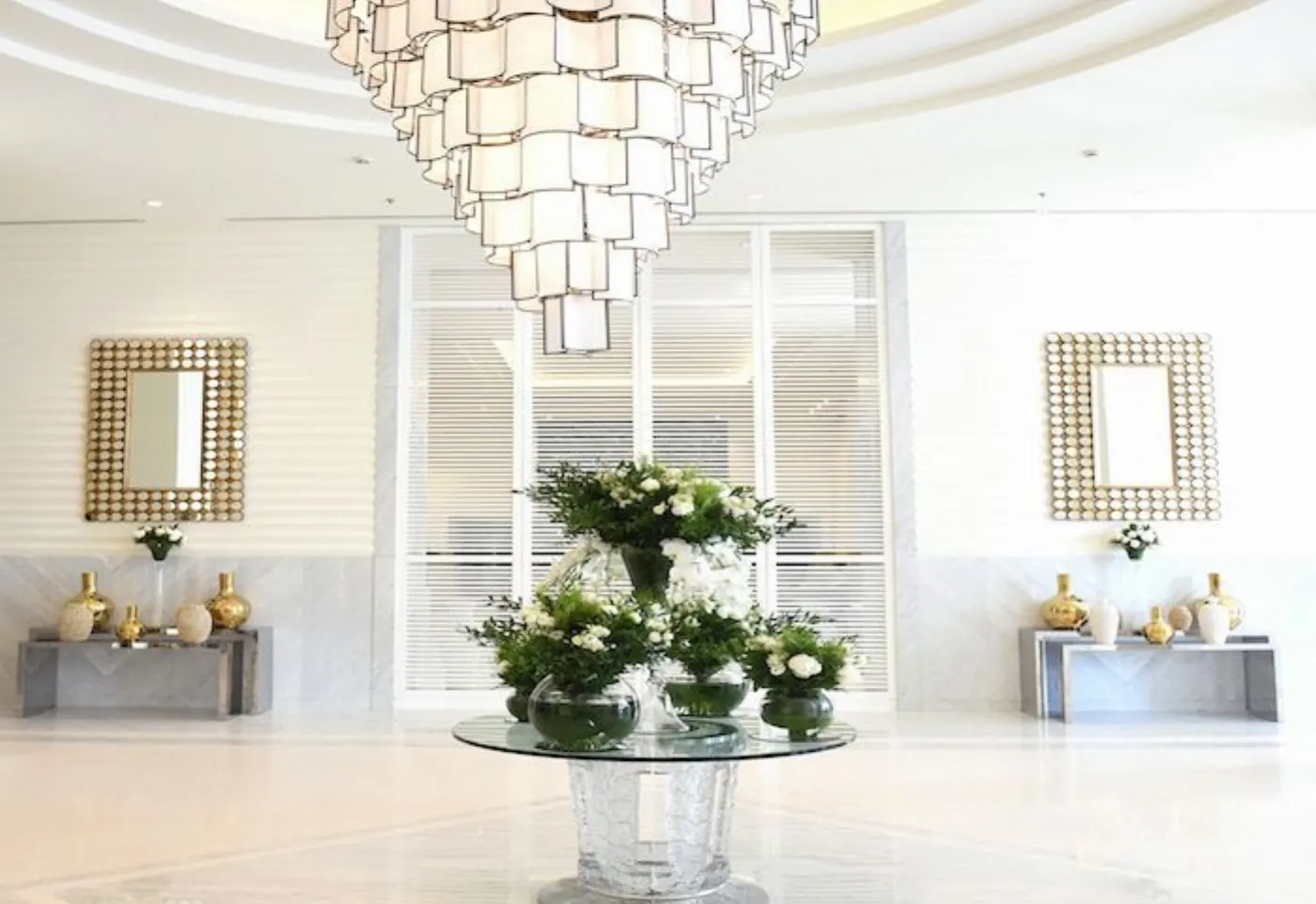 Hall d'hôtel très clair, lustre à abat-jour blancs, consoles garnies de vases dorés et composition florale blanche