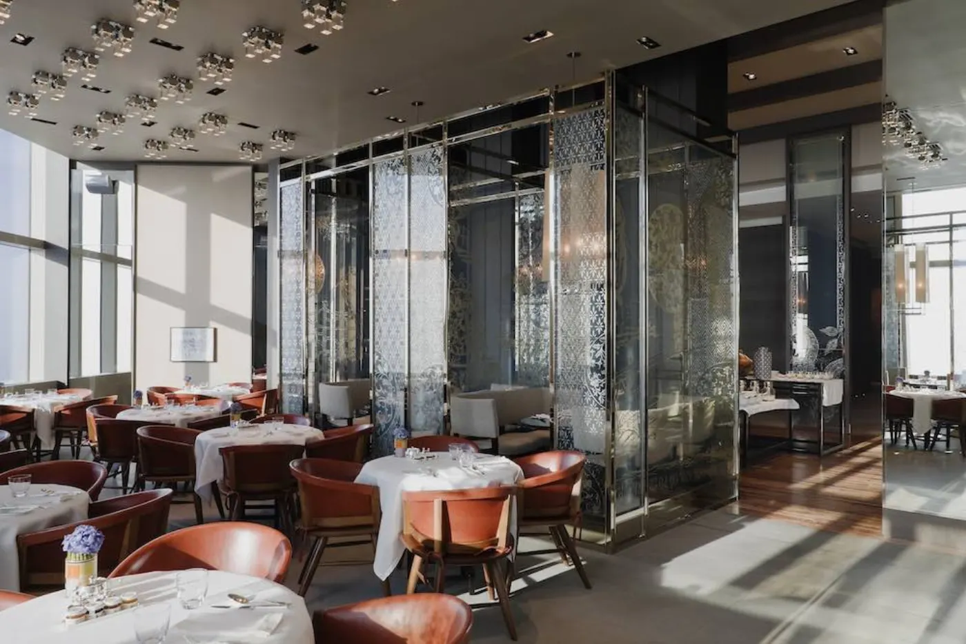 Salle de restaurant baignée de lumière, tables rondes nappées de blanc, fauteuils cuir orangé et paravents de verre gravé