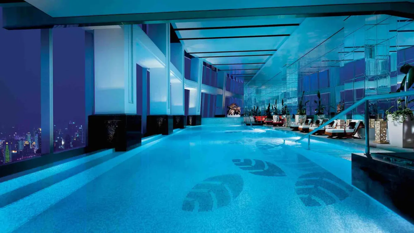 Piscine intérieure éclairée en bleu au sommet d'une tour, transats alignés et lumières de la ville la nuit