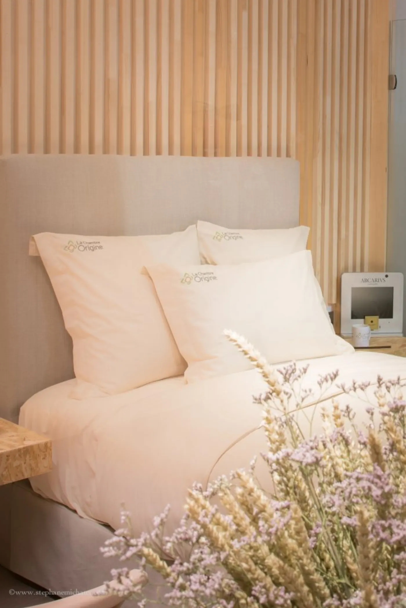Lit aux coussins brodés La Chambre Origine devant un mur de tasseaux clairs, fleurs séchées au premier plan