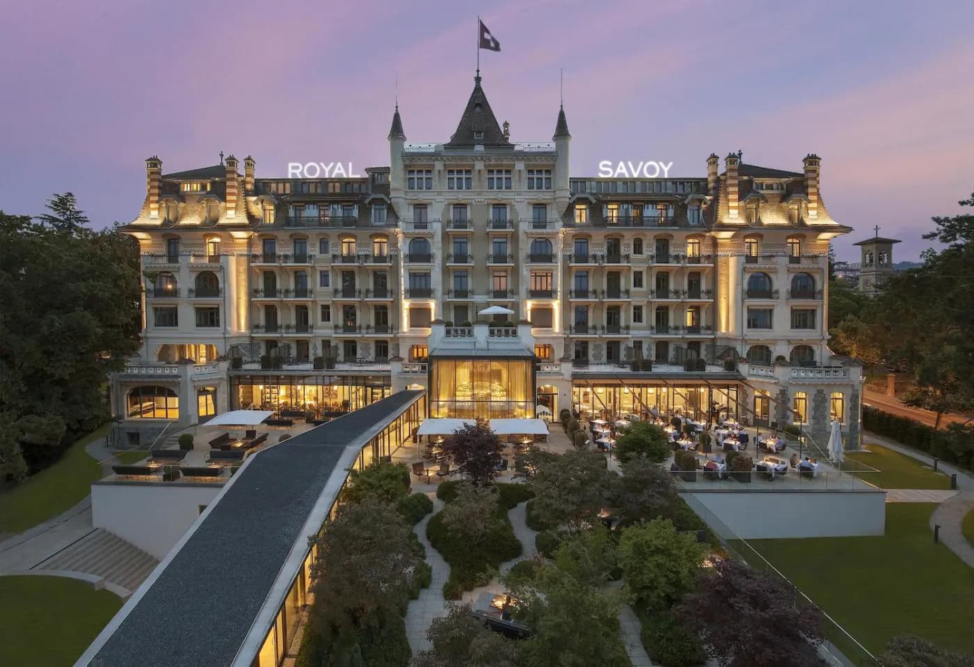 Le Royal Savoy illuminé au crépuscule, enseigne sur le toit, drapeau suisse et terrasse restaurant côté jardin