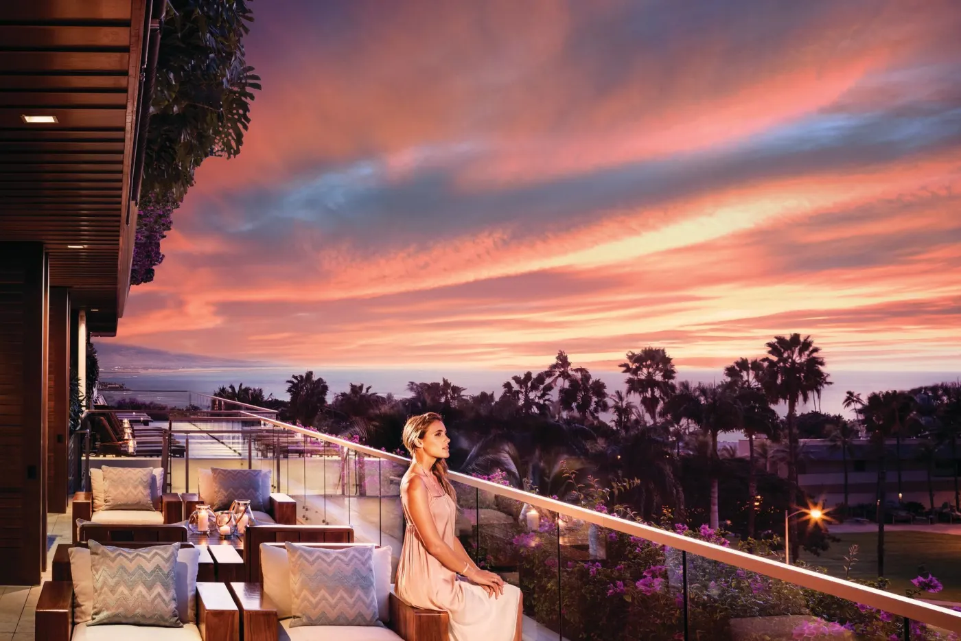 Femme en robe rose assise sur une terrasse lounge face aux palmiers et à l'océan sous un ciel de coucher de soleil