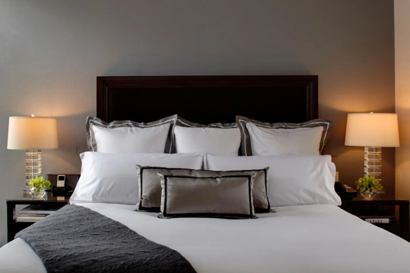 Lit d'hôtel à tête de bois foncé, oreillers blancs bordés de gris et lampes de chevet allumées de part et d'autre
