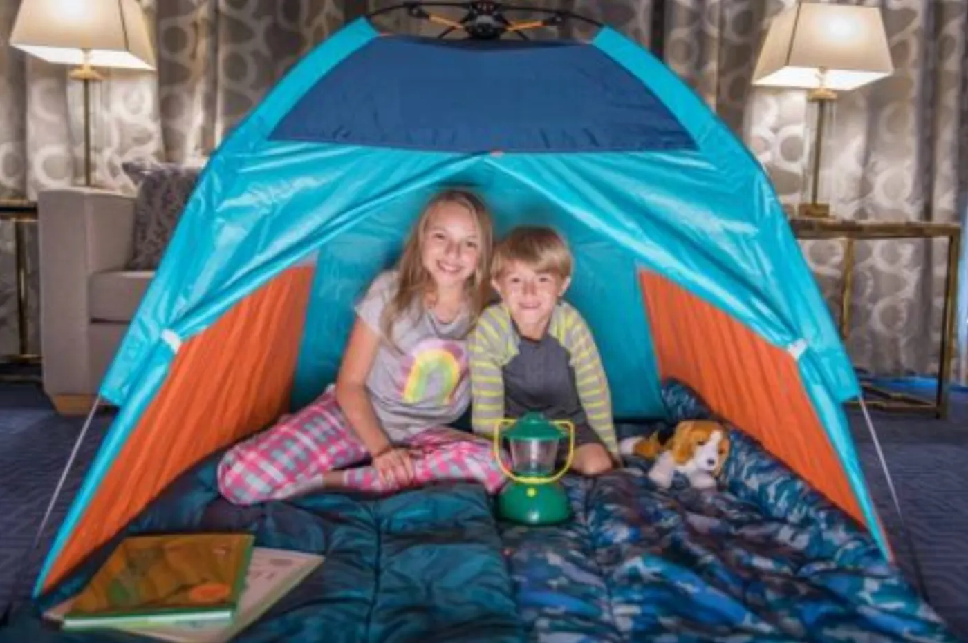 Deux enfants souriants installés dans une tente de camping montée à l'intérieur d'une chambre d'hôtel