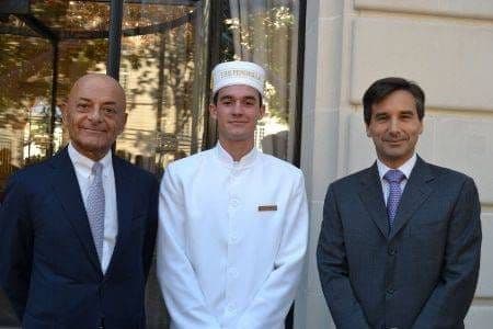 Deux dirigeants du Peninsula encadrant un jeune chasseur en uniforme blanc devant l'entrée de l'hôtel