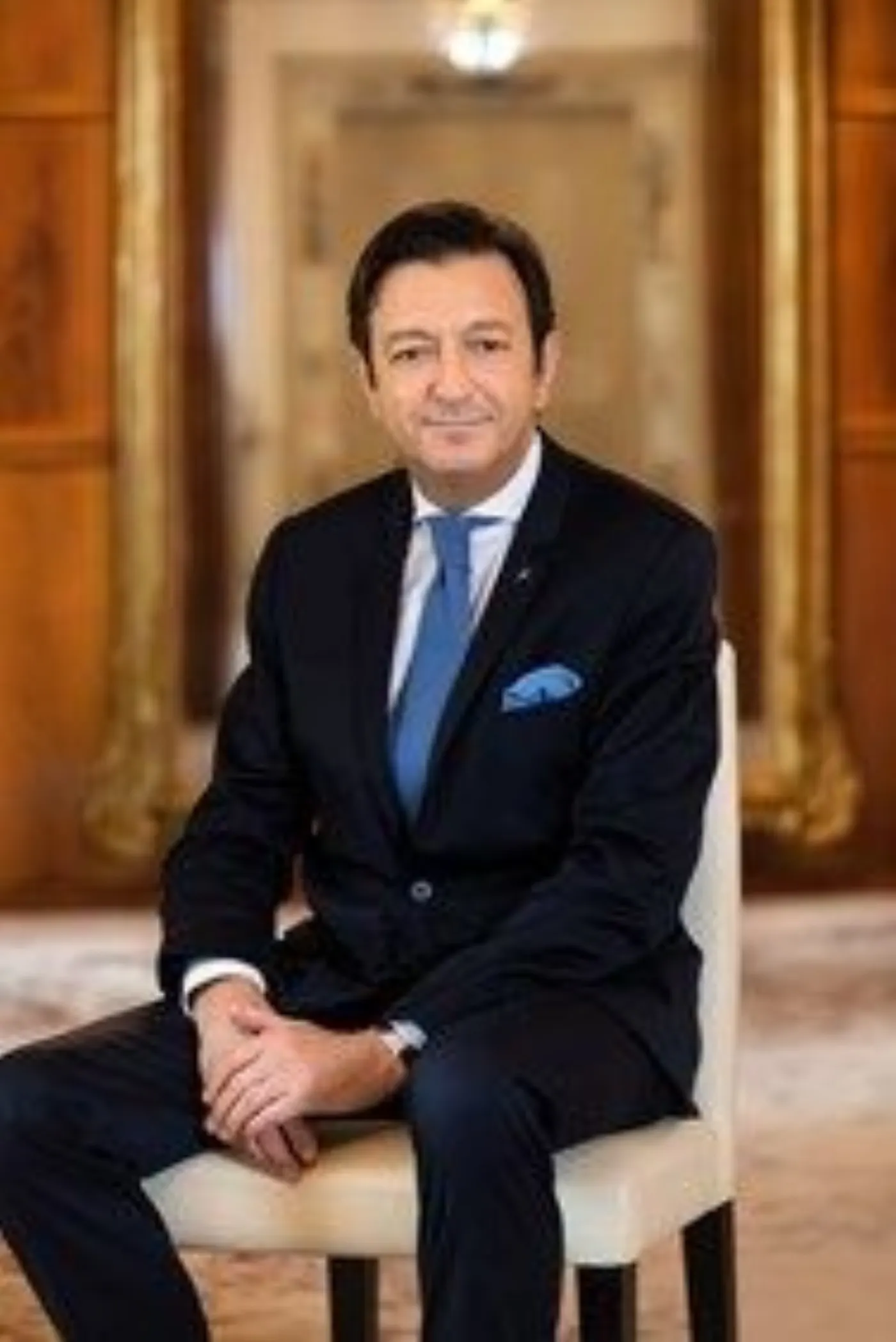 Homme en costume sombre et cravate bleue, assis sur une chaise blanche dans un salon aux boiseries dorées