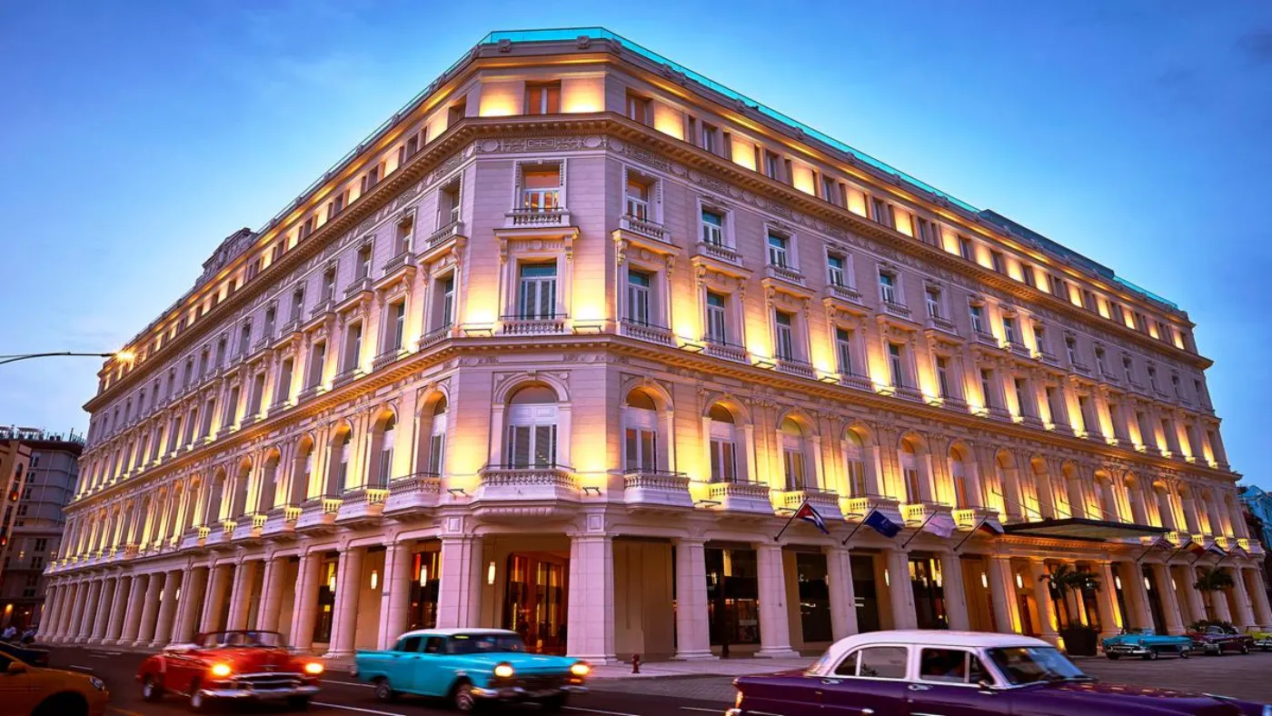 Façade néoclassique d'un hôtel illuminée au crépuscule, voitures américaines vintage passant devant