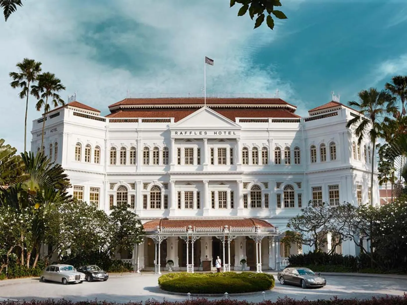 Façade blanche coloniale du Raffles Hotel Singapour, toit de tuiles rouges et palmiers alentour