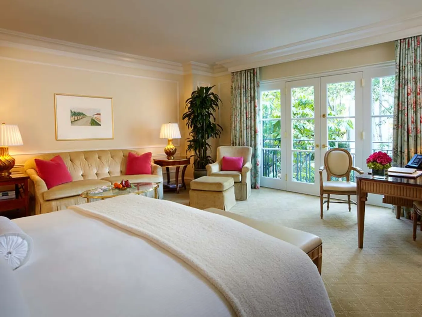 Chambre aux tons crème avec canapé capitonné, coussins roses et portes-fenêtres ouvrant sur la verdure