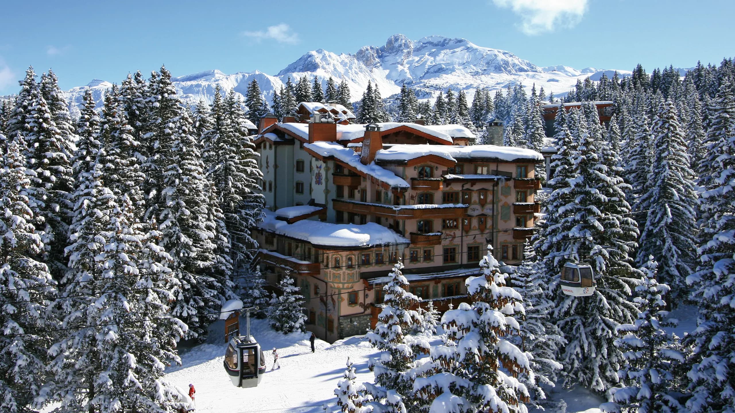 Hôtel de montagne aux façades peintes, cerné de sapins enneigés et survolé par une télécabine