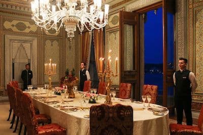 Salle à manger de palace dressée aux chandelles, trois maîtres d'hôtel en attente sous le lustre de cristal
