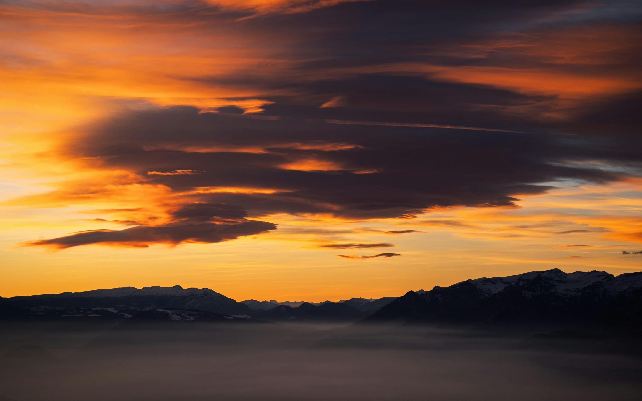 Coucher de soleil orangé sur une mer de nuages, silhouettes de sommets enneigés à l'horizon