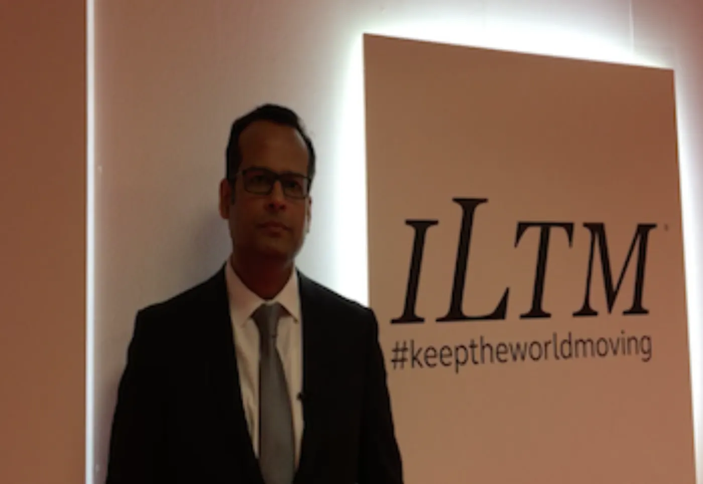 Homme en costume sombre et lunettes devant un panneau ILTM portant le mot-clic keeptheworldmoving