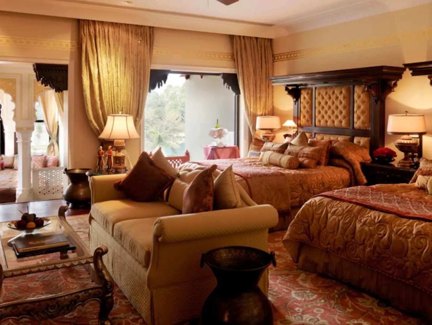 Suite indienne aux tons ocre avec deux lits à tête sculptée, canapé beige, rideaux dorés et tapis à motifs