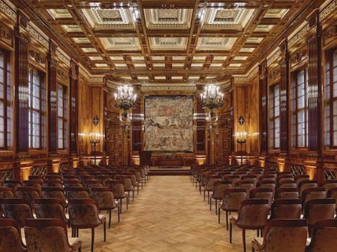 Grande salle boisée à plafond à caissons dorés, lustres et fauteuils alignés face à une tapisserie
