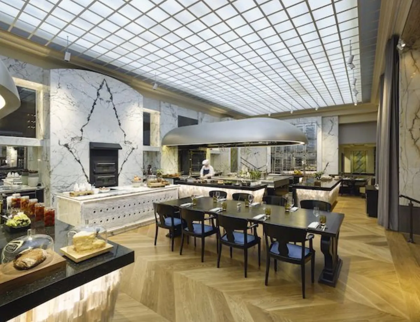 Restaurant sous verrière avec cuisine ouverte, un chef au piano, buffet de pains et fromages, parquet à chevrons