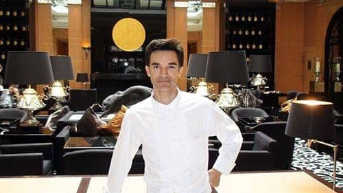 Chef in white jacket standing in a hotel lounge, black lampshades and a golden disc behind him