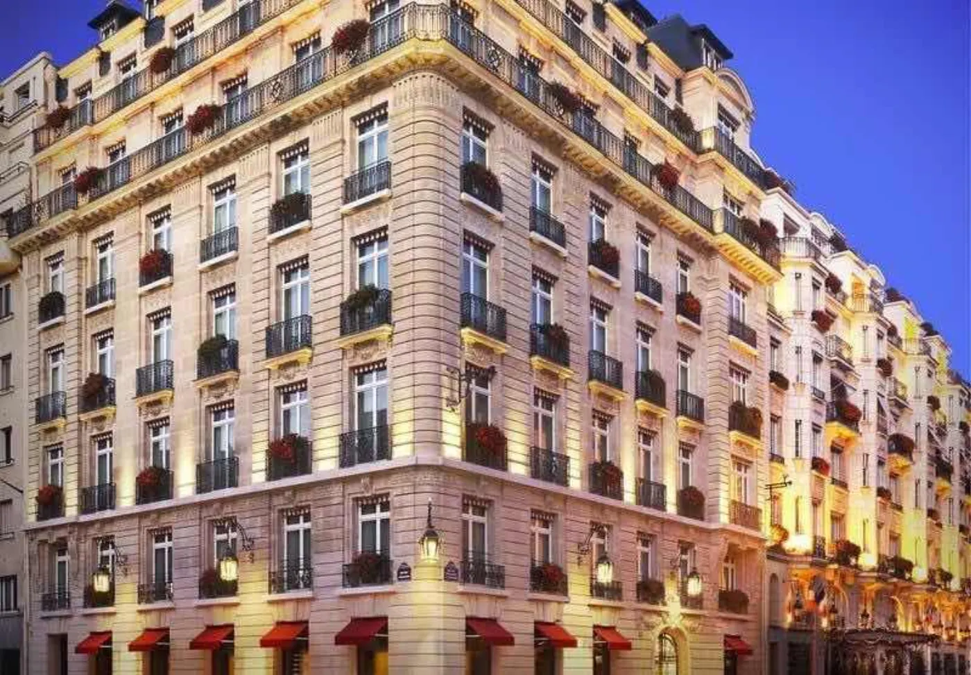 Façade haussmannienne d'un hôtel parisien illuminée au crépuscule, stores rouges et balcons fleuris