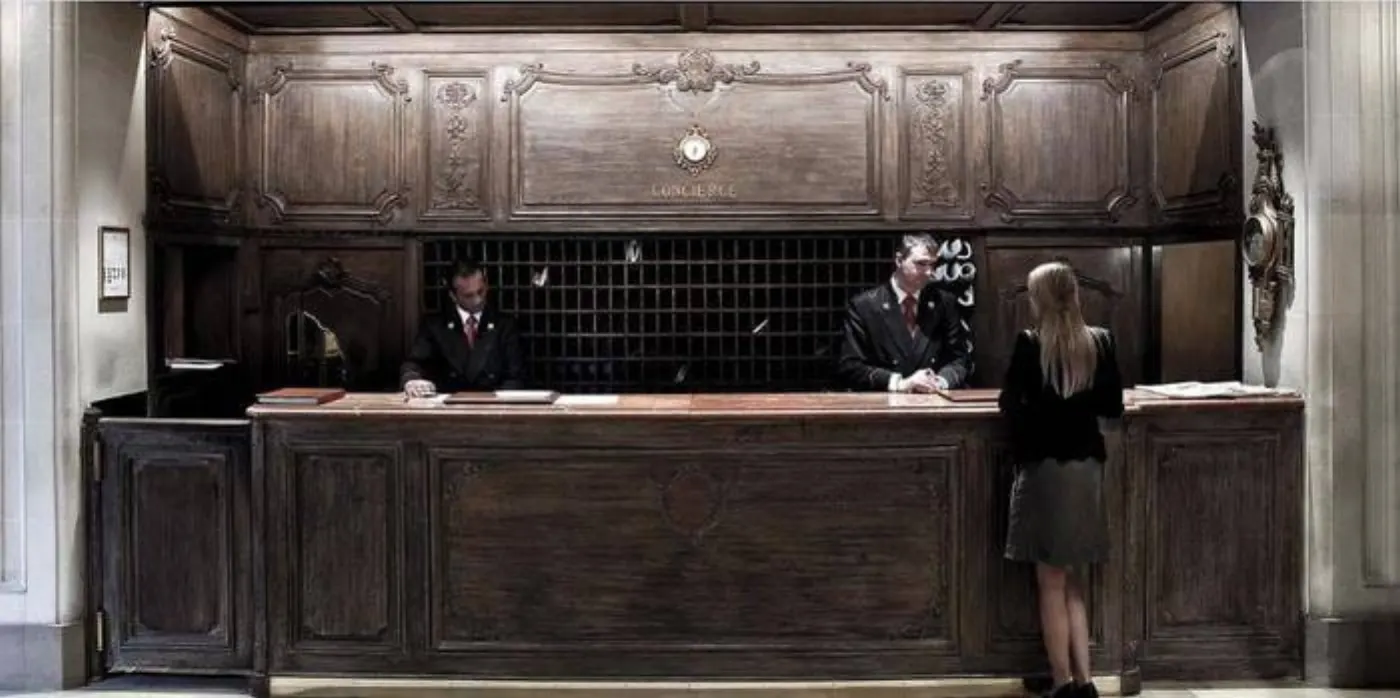 Comptoir de conciergerie en bois sculpté, deux concierges en uniforme et une cliente devant le casier à clés
