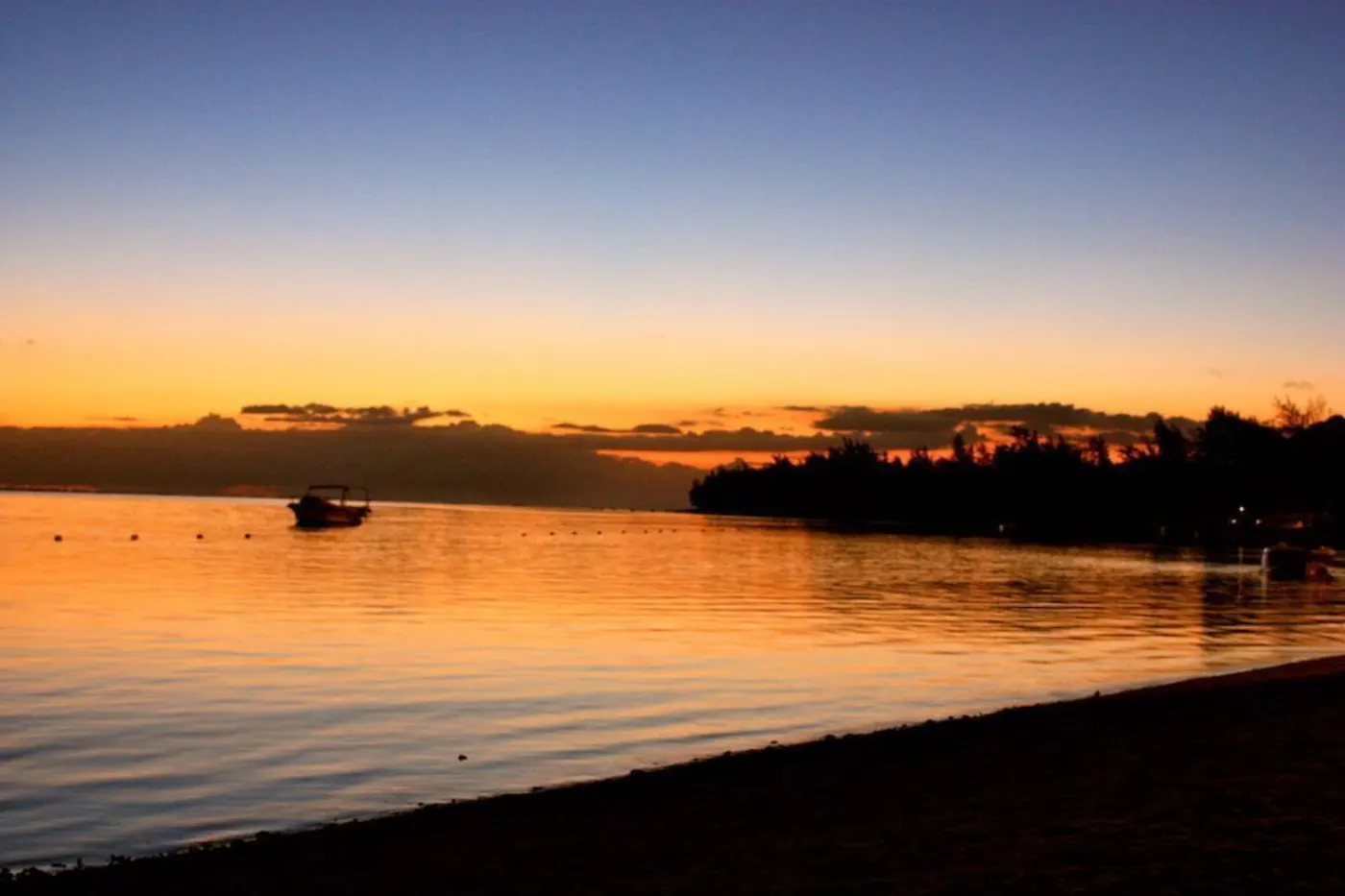 Coucher de soleil orangé sur un lagon, bateau à l'ancre et rivage boisé en silhouette