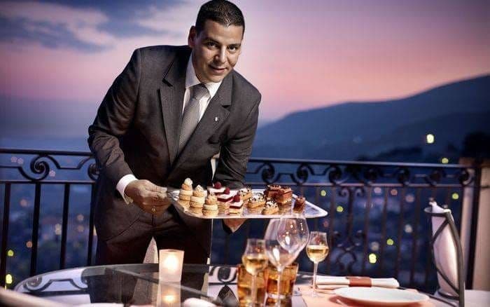 Maître d'hôtel présentant un plateau de mignardises sur une terrasse au crépuscule, la baie en contrebas
