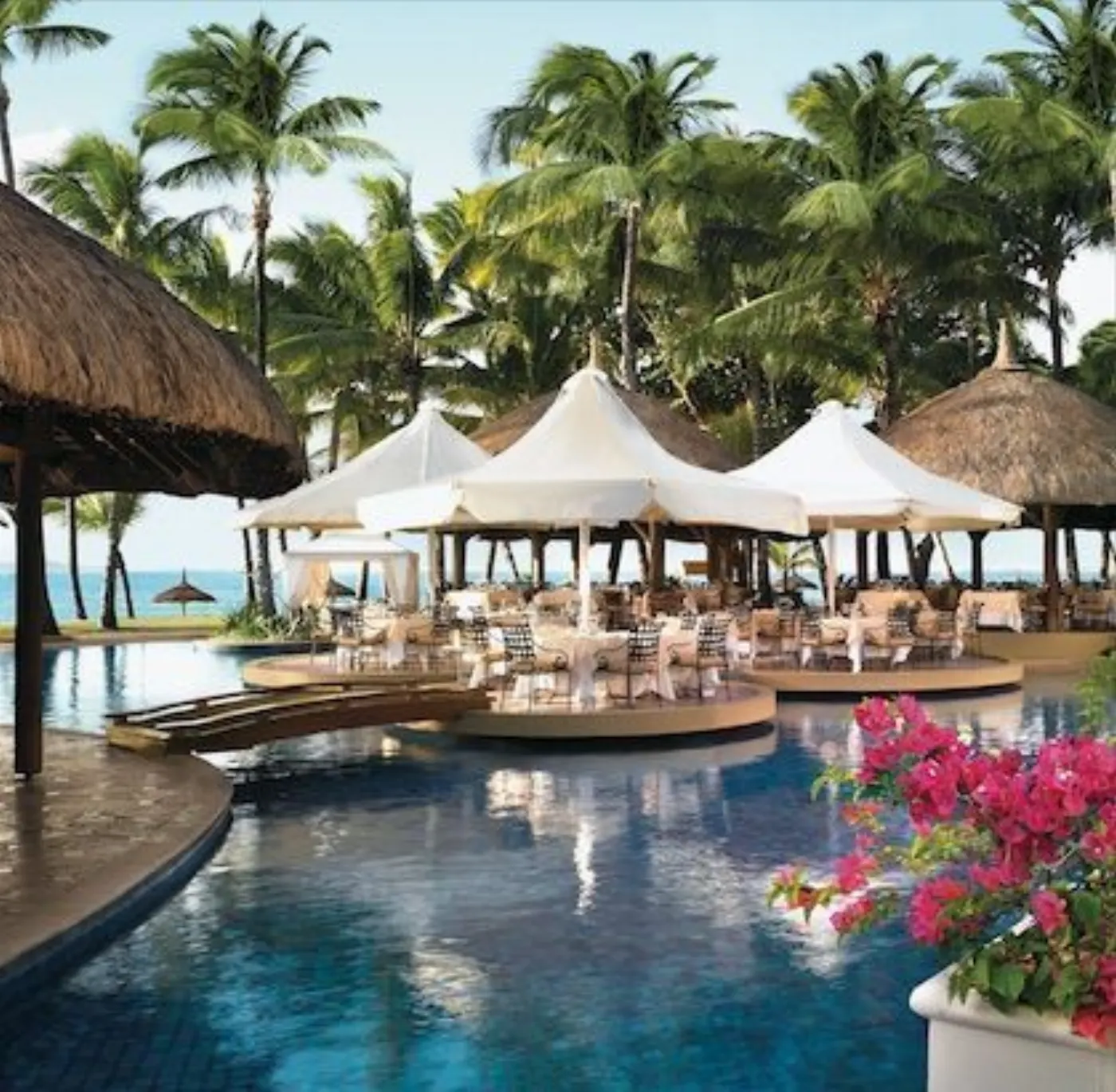Restaurant dressé sur des îlots au milieu d'une piscine, sous parasols blancs et toits de chaume, entre les palmiers