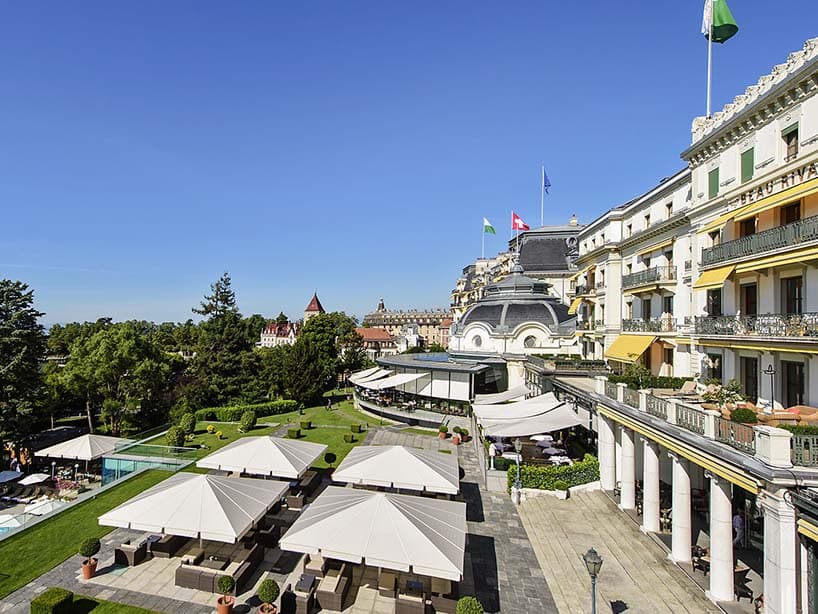 Terrasse du Beau-Rivage Palace : parasols blancs, pelouses taillées et façade à balcons sous le ciel bleu