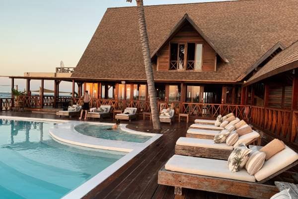 Beachfront resort at dusk: pool, cushioned day beds and a wooden lodge facing the sea