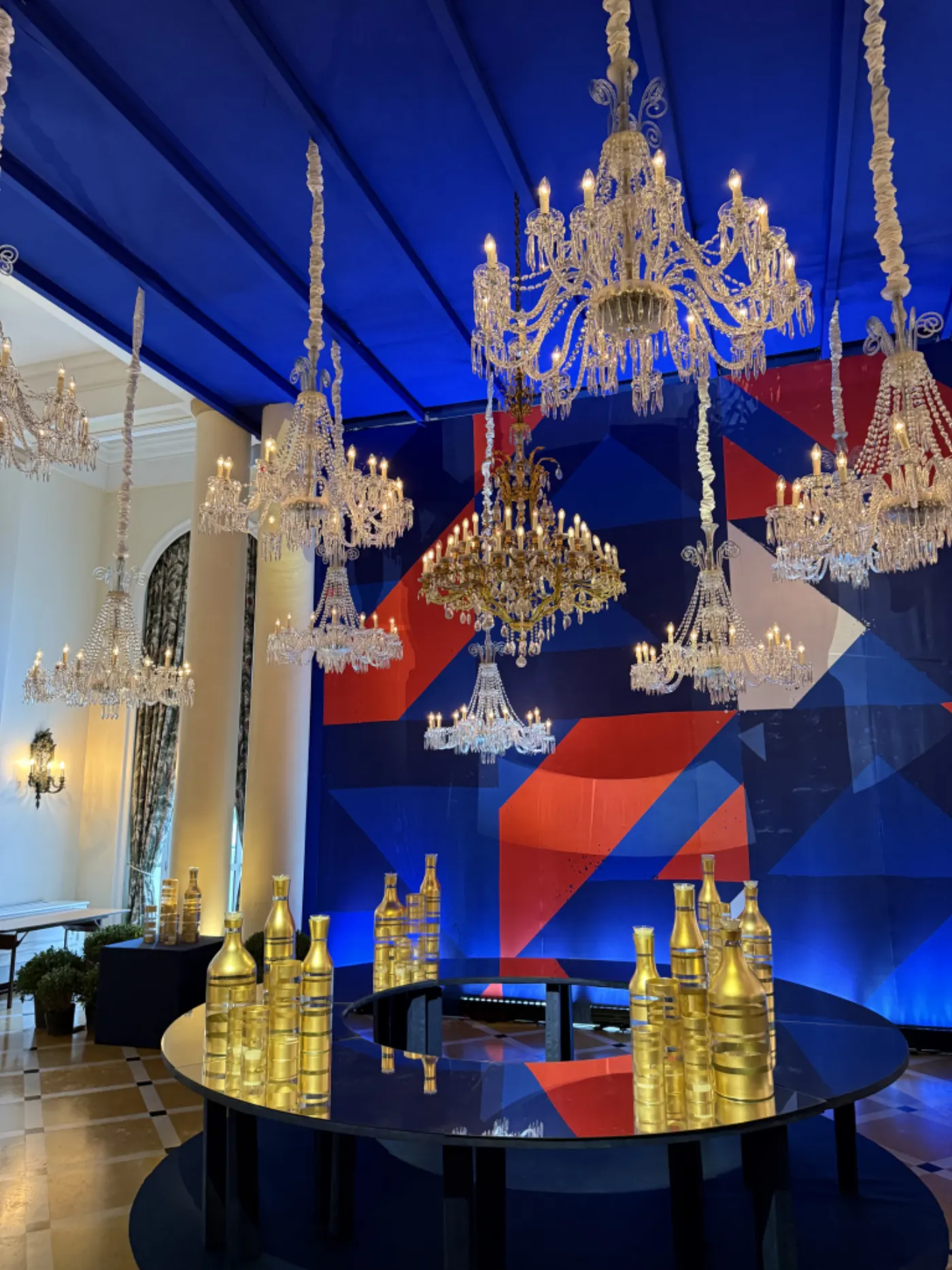 Salon d'hôtel décoré pour une soirée : lustres en cristal, plafond bleu tendu et bouteilles dorées sur table ronde