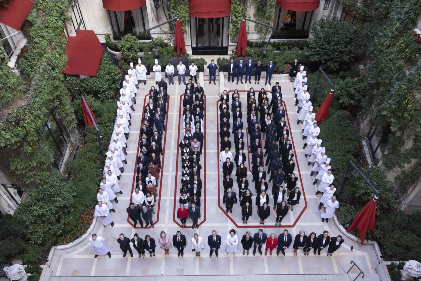 Personnel en tenue et chefs en veste blanche alignés dans la cour intérieure du Plaza Athénée, stores et parasols rouges