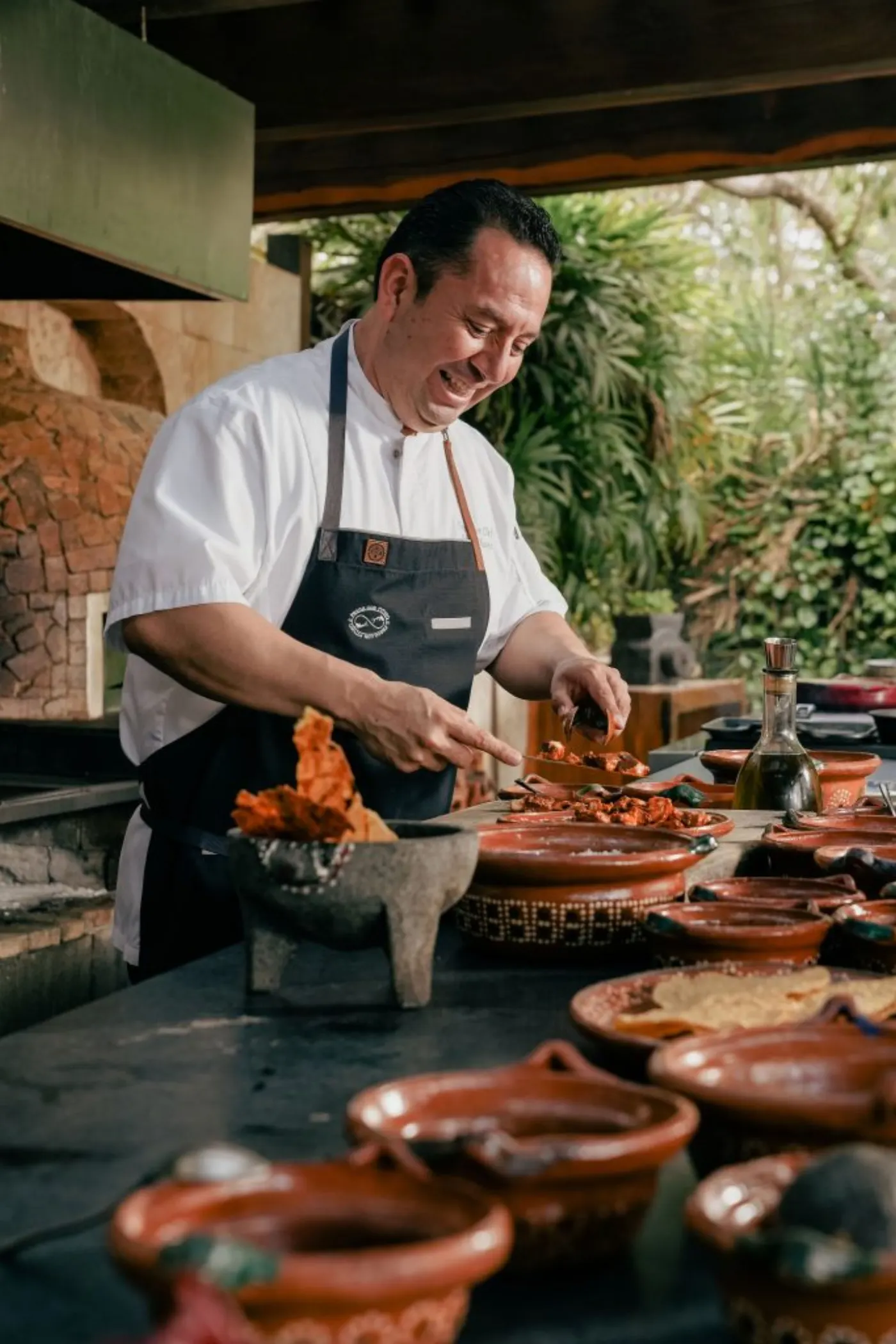 Le chef Edgar Chávez, en tablier de jean, dresse des viandes grillées près d'un four à bois et de plats en terre cuite