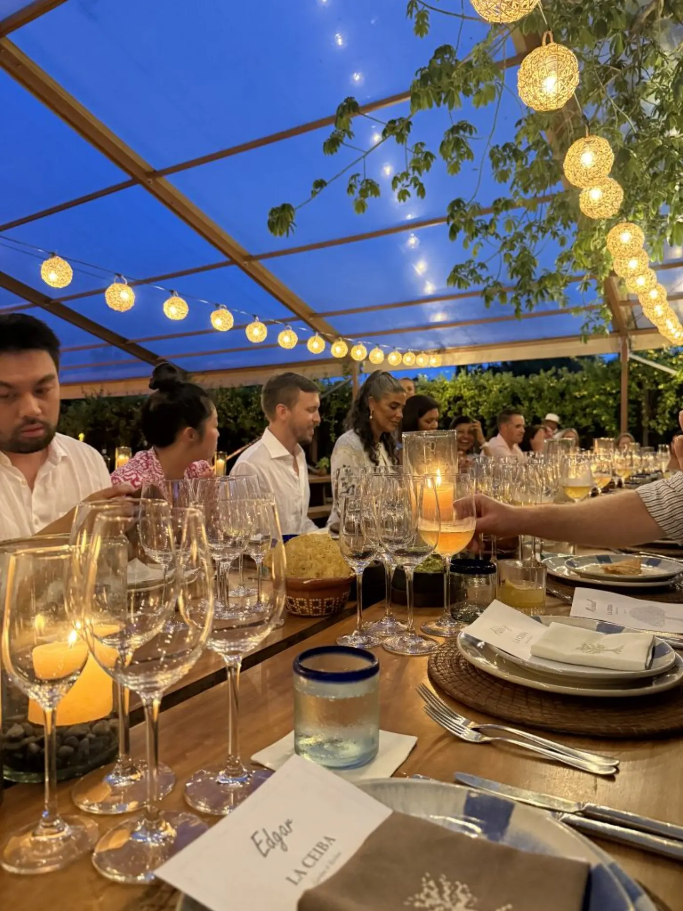 Dîner sous une tente au crépuscule, longue table de verres et bougies, guirlandes lumineuses et carton Edgar La Ceiba