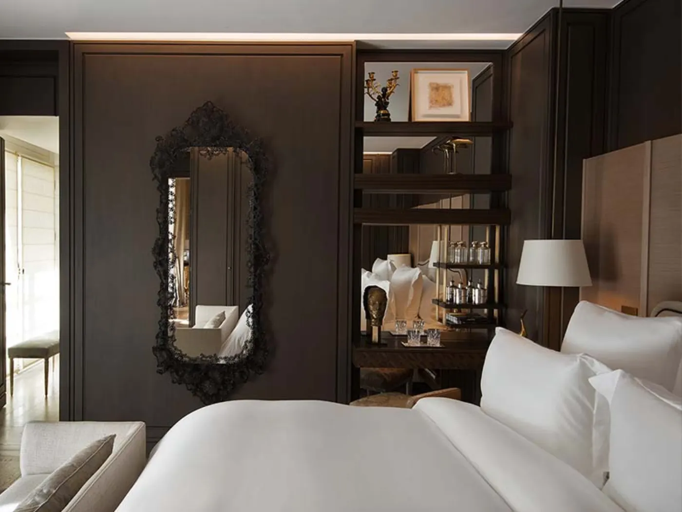Chambre aux boiseries sombres, grand miroir baroque noir, lit blanc et minibar dressé sur des étagères éclairées