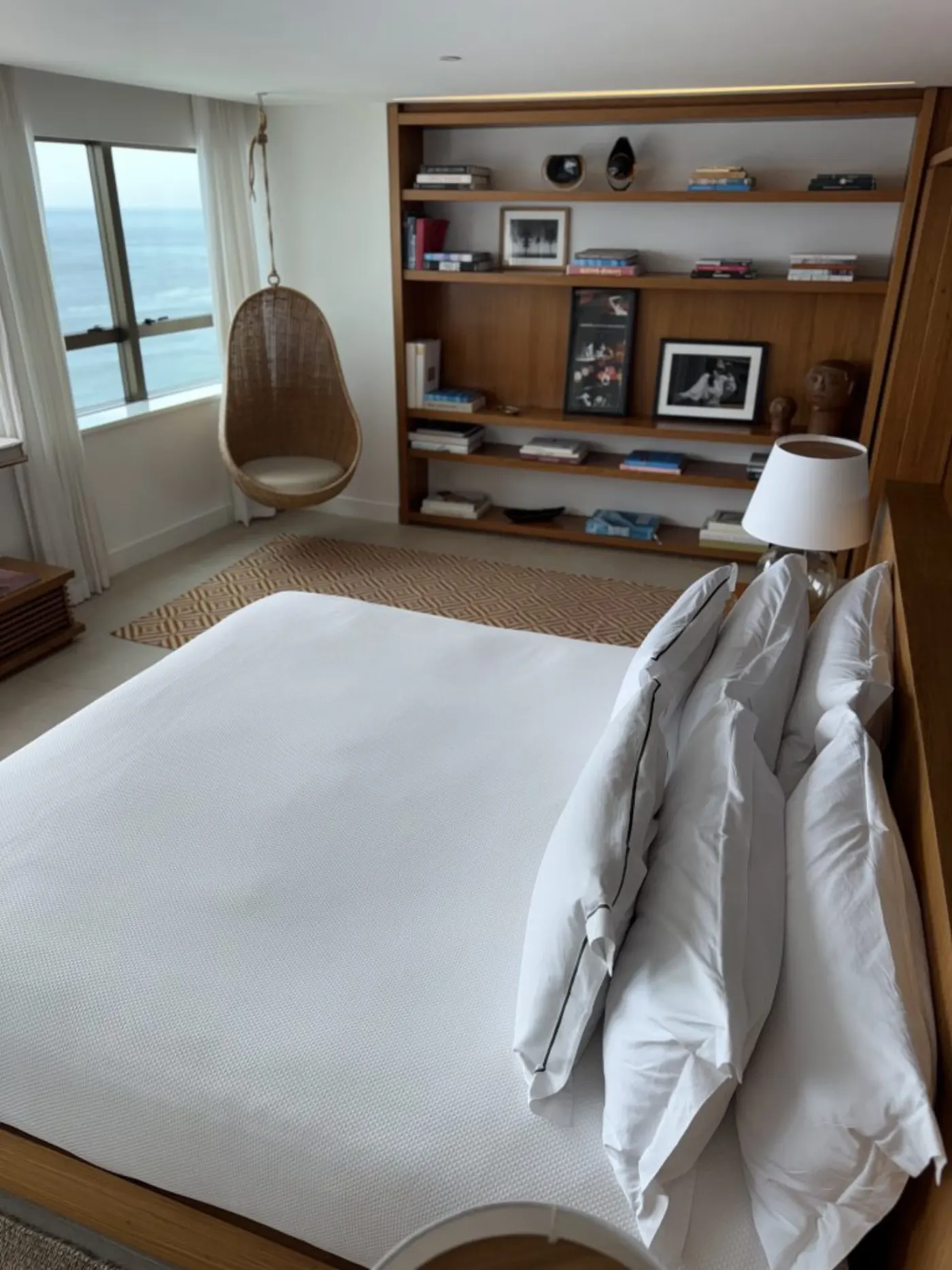 Chambre face à l'océan avec fauteuil suspendu en rotin, bibliothèque en bois et grand lit blanc