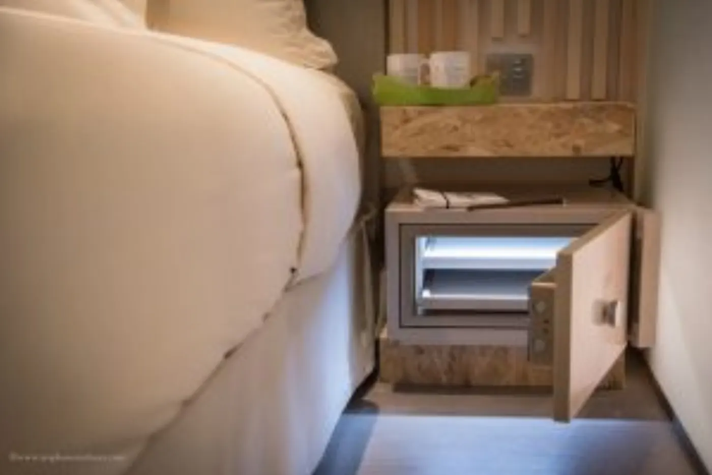 Table de chevet en bois OSB avec coffre-fort ouvert et éclairé, à côté d'un lit à la couette blanche