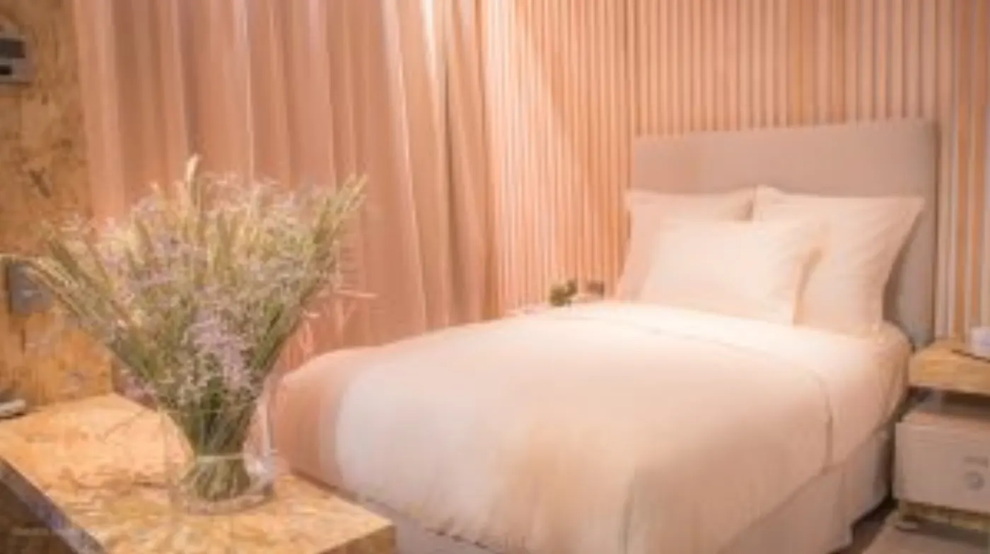 Chambre baignée d'une lumière rosée, lit blanc devant un mur cannelé et bouquet de fleurs séchées