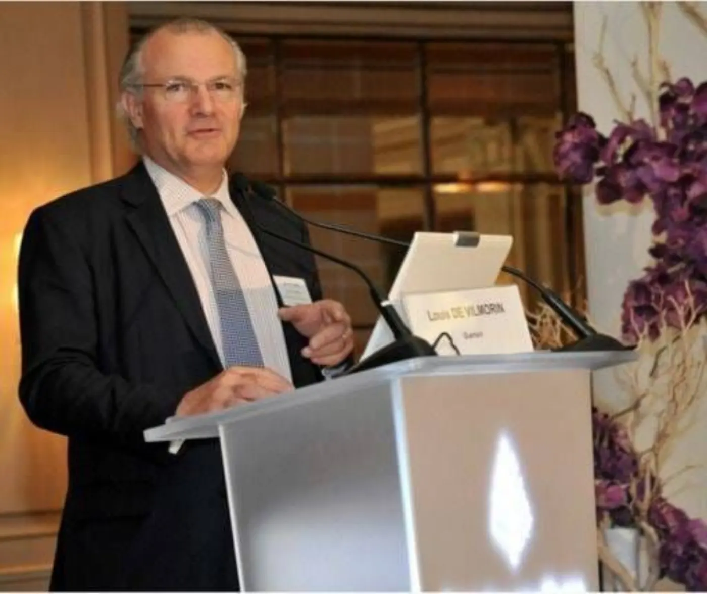 Homme en costume sombre s'exprimant à un pupitre, chevalet au nom de Louis de Vilmorin, orchidées violettes à droite