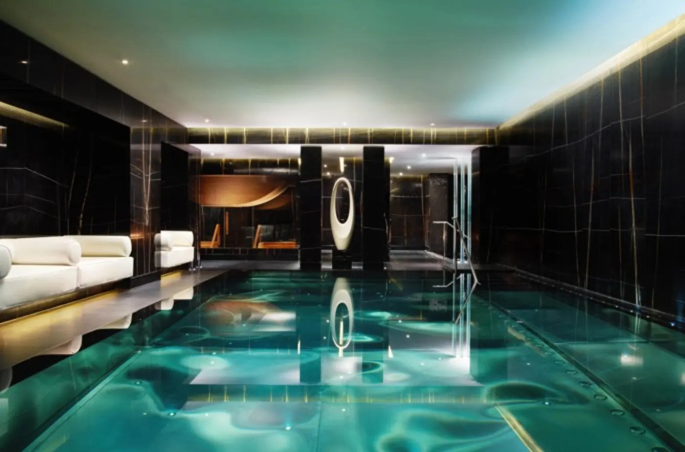 Piscine intérieure de spa aux murs de marbre noir, eau turquoise éclairée et banquettes blanches